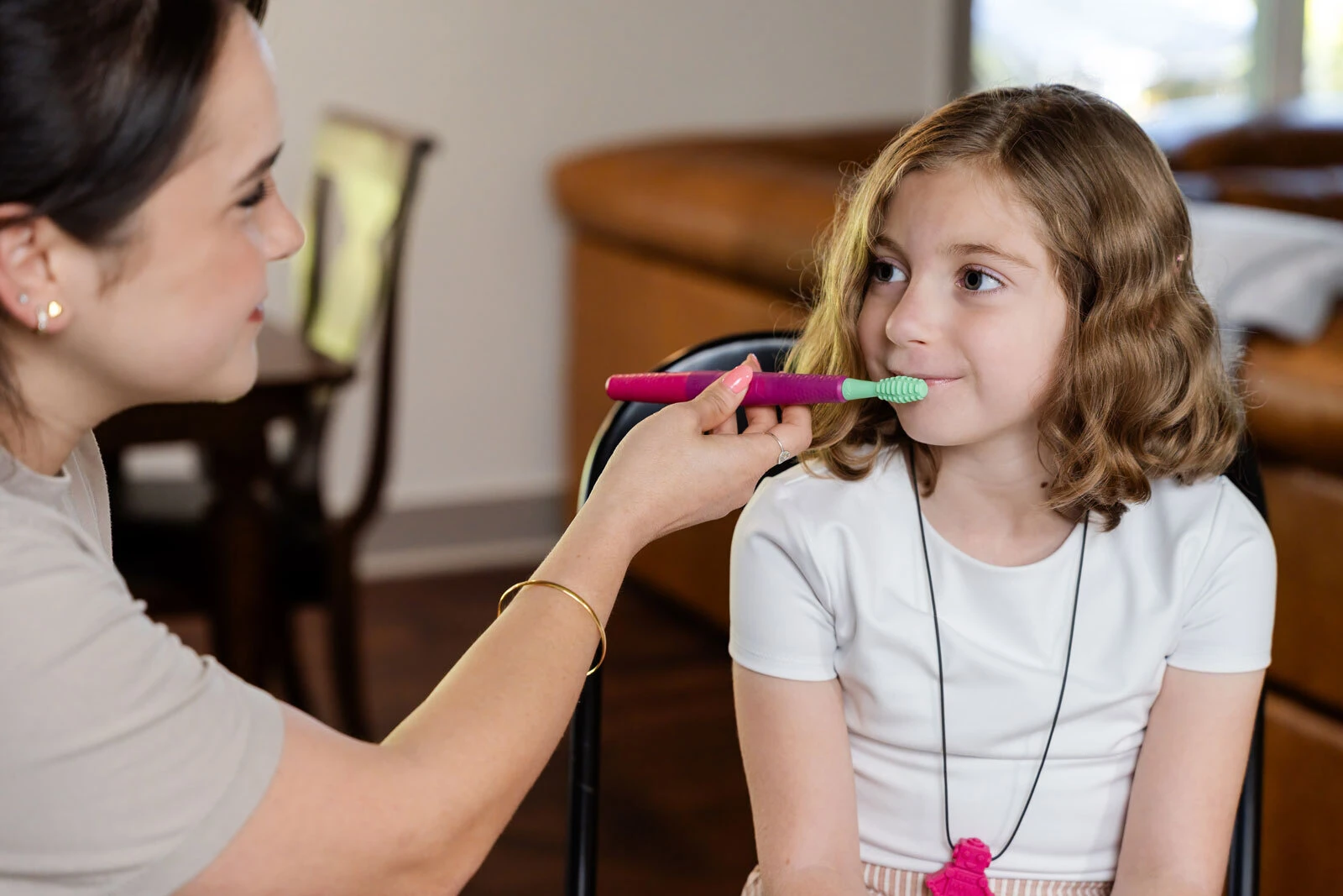 A clinician gently uses the Z-Vibe Plus with a smiling school-age girl who is wearing a chew necklace.
