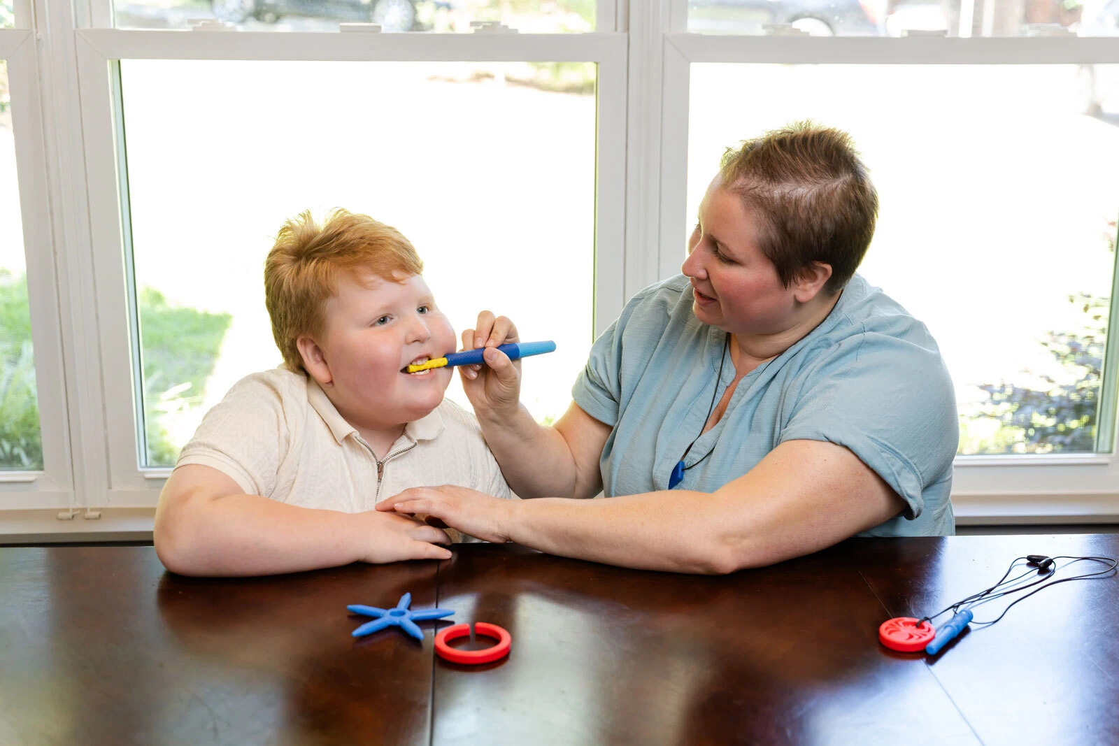 A smiling boy uses the Z-Vibe Plus with a caregiver, with several ARK tips and a chew necklace on the table.