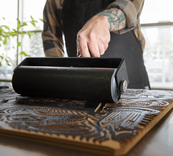 Close-up of a hand rolling ink with a large brayer over a carved wooden block.