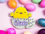 A personalized enamel pin featuring a cheerful yellow chick hatching from an egg. The chick has a cute expression with closed eyes and a big smile. The lower half of the egg shell contains the name 'Andrea' in a playful, handwritten font with pink highlights. The year '2021' is written at the bottom of the shell. The pin is set against a pink background, suggesting a fun and custom-made accessory.