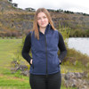 A woman stands by a lake wearing a navy classic softshell vest, with her hands in the pockets and a scenic background.