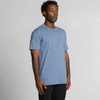 A model wears a light blue T-shirt with a relaxed fit. The shirt features a simple design and includes a logo.