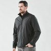 A man wearing a black softshell jacket with a zipped front and high collar, featuring a logo.
