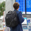 A person carrying a black backpack stands in an airport near a flight information board. The backpack has a logo.