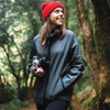 A woman wearing a dark green hooded rain jacket and a red beanie, holding a camera in a forest setting.