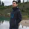 A man stands by a body of water, wearing a black softshell jacket with blue zippers in a natural setting.