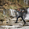 A man in a grey rain jacket jumps over a rocky stream, with a waterfall in the background. The jacket features a logo.