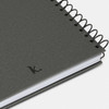 A grey spiral-bound journal with a textured cover and a simple black logo on the bottom corner.