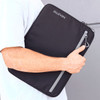A laptop sleeve in black with a grey zipper, featuring a logo, held under someone's arm.