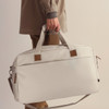 A duffle bag in light grey, being carried by a person with a neutral outfit, featuring a logo and leather accents.