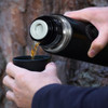 A black 750ml vacuum flask is being poured into a black cup, surrounded by a natural background.