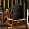 A black backpack sits on a wooden chair beside a speaker system and potted plant. It features multiple compartments and a logo.