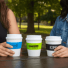 Three small cups in white, each with a different coloured band: blue, green, and black, held by two people.