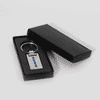 A silver keychain in a black presentation box, featuring a simple design and a logo.