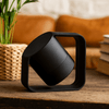 A black wireless speaker with a unique geometric design, set against a decorative background with plants and books.