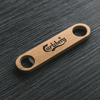 A bamboo bottle opener in a natural wood colour, featuring a logo and two openings.