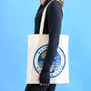 A cream-coloured tote bag with a logo, held by a person against a blue background.