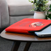 A red laptop sleeve featuring a logo, resting on a wooden table beside a plant and a laptop.