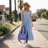 A woman wearing a blue dress carries a purple tote bag with a logo while walking on a sunny street lined with palm trees.