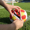 A football being zipped into a foldable gym bag, featuring red and yellow panels on the ball. The scene is on a grassy field.