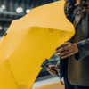 A yellow BLUNT Metro Umbrella is held by a person wearing a dark outfit, showcasing its sleek design.