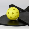 A yellow pickleball with holes, resting on a black paddle and a round black surface, featuring a logo.