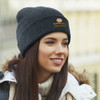 A dark grey beanie worn by a woman, featuring a logo on the front and set against an outdoor backdrop.