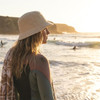 A woman stands by the ocean in a beige bucket hat, with surfers visible in the background during sunset.