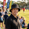 A man wearing a black Leroy Cord Bucket Hat smiles at a sports field, with people and tents in the background.