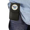 A black phone pouch with a belt clip, featuring a circular logo on the front, attached to a person’s waist.