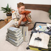 A set of grey travel packing cubes arranged beside a suitcase, with a man sitting on a bed, holding an orange item.