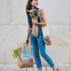 A woman in blue jeans carries an iridescent rainbow PVC bag, flowers, and a net bag with fruit.