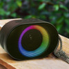 A compact black Bluetooth speaker with an LED light ring displaying rainbow colours. It features a textured surface and a carry strap.