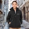 A woman wearing a black softshell jacket with a hood, featuring a logo on the chest, stands in a cobblestone alley.