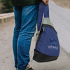 A slinger bag in navy blue with grey and black accents, featuring a logo. It is being held by a person on a path.