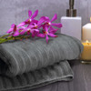 A grey bath towel folded neatly with a bouquet of purple orchids, a white container, and a candle in the background.