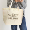 A natural-coloured tote bag with a logo, held by a person wearing a grey sweater and jeans.