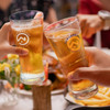 Two clear glass beer glasses filled with golden beer, toasting over a dining table with food. Each glass has a logo.