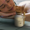 A glass jar containing white bath salts, topped with a cork lid and wooden spoon, beside a person relaxing in a bath.