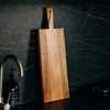 A wooden chopping board with a handle, featuring rich brown tones, placed on a dark textured surface.