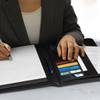 A sleek black portfolio with a logo, held by hands, on a wooden surface with other documents nearby.