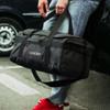 A black duffle bag with a logo, held by a person in casual jeans and red sneakers.