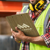 A bamboo clipboard with a logo, held by a person wearing a high-visibility vest and earmuffs in an industrial setting.