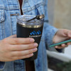 A drink bottle featuring a black exterior and a transparent lid, held by a person with a denim jacket. It has a logo.