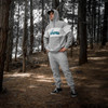 A unisex hoodie in grey with a logo, displayed on a person standing amidst trees in a forest setting.