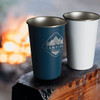 A drink bottle set featuring a blue and a white tumbler, both with a logo, set against a campfire background.
