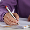 A green recycled plastic pen with a red cap, held by a hand, writing in a notepad. The pen features a logo.