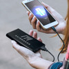 A black power bank is being held while charging a smartphone. The power bank has a logo on it.