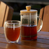 A transparent coffee plunger with a wooden lid beside a clear glass cup filled with brown liquid, set on a wooden table.