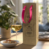 A champagne ribbon handle paper bag in natural brown with pink handles, beside a glass of white wine and snacks.